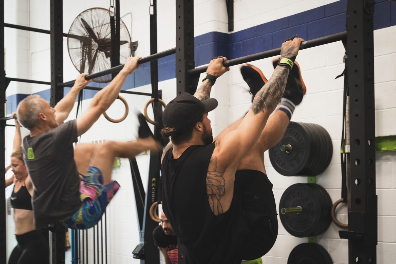 Two men are performing pull-ups at CrossFit Haapu in Nerang, Gold Coast, lifting their legs toward the bar as part of their CrossFit programs. The man in the foreground has tattoos and is wearing a black tank top and hat. The man in the background is wearing a gray shirt and colorful shorts. Weight plates are visible on the wall behind them.